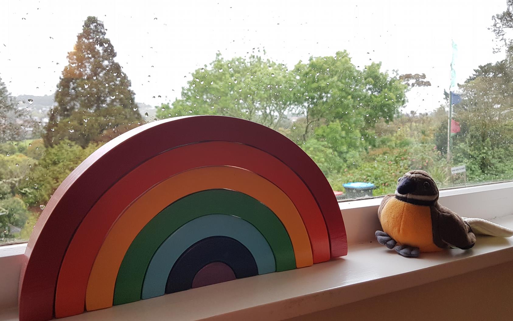 A wooden rainbow and a pīwakawaka (fantail) soft toy sit on a windowsill. It's rainy outside